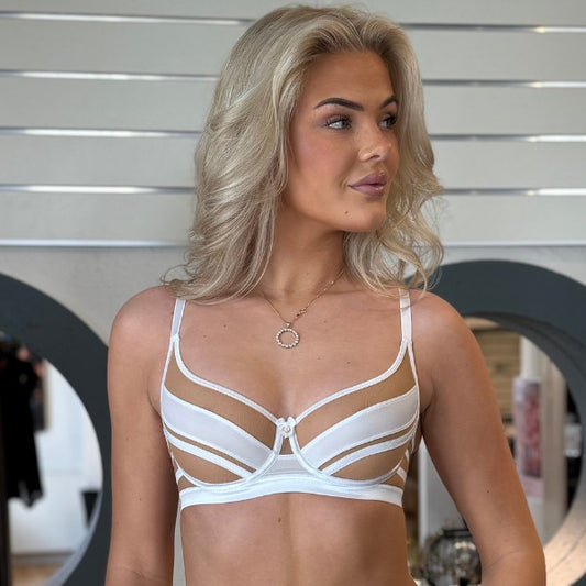 En kvinde med langt, bølget blond hår viser en hvid Axami-bh med stropper og transparente paneler for luksus og komfort. Indendørs står hun ved siden af hvide paneler og cirkulære spejle og kigger til siden, mens hun bærer en delikat halskæde med vedhæng.