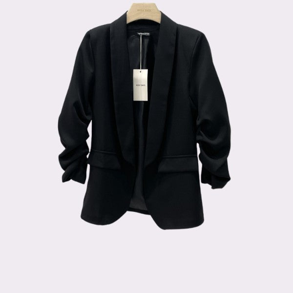 Rosy Days Blazeren er en sort skræddersyet blazer med rynkede ærmer, hakkrave, to lommer med klap foran og et hvidt mærke ved kraven. Denne elegante og tidløse favorit fra Rosy Days hænger på en træbøjle mod en lys baggrund.