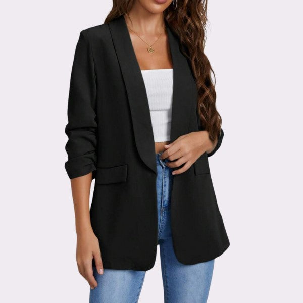 En kvinde bærer den elegante, tidløse Rosy Days-blazer over en hvid ribbet tubetop og blå jeans. Med opskubbede ærmer og bølget brunt hår pryder hun med en gylden hjertehalskæde. Hendes hånd hviler på taljen mod en lysegrå baggrund.