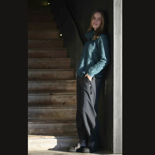 En kvinde med langt hår, klædt i en blågrøn E Avantgarde-sweater, løse sorte bukser og sorte støvler, læner sig afslappet op ad en solbeskinnet grå væg ved en trætrappe og kigger ind i kameraet med et lille smil.