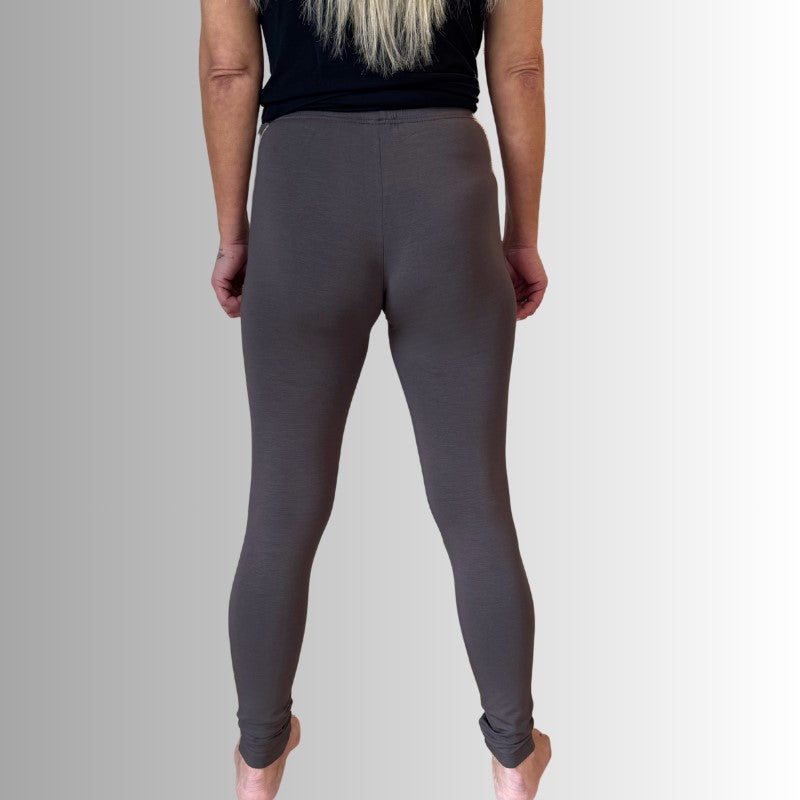 En person med langt, lysblondt hår bærer en sort top og tætsiddende leggings fra E Avantgarde, står barfodet og kigger væk mod en grå baggrund med armene langs siden.