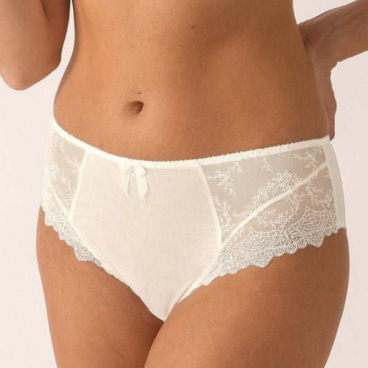 Et nærbillede af en kvinde iført cremefarvede blondetrusser fra Empreinte med transparent blomsterbroderi og en lille sløjfe i taljen, sat op mod en neutral baggrund. Kun hendes torso og overlår er synlige.