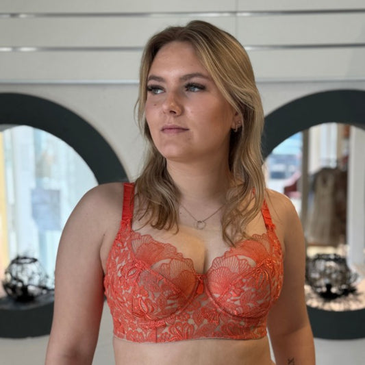 En kvinde med langt blond hår bærer en orange Empreinte-bh af blonder, der er fremstillet med fransk elegance og støtte, kombineret med en halskæde. Hun står indendørs foran en væg med cirkulære udskæringer og stirrer let til siden med et neutralt udtryk.