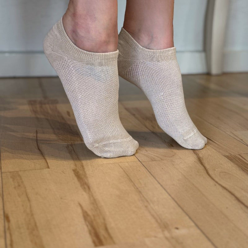 En person står på tæer på et trægulv iført lysbeige, nedringede fodsko fra Festival Strømper. De enkle strikkede sokker er lavet af blød bambusfiber til svedtransport og vises med en hvid væg og en lys stol i baggrunden.