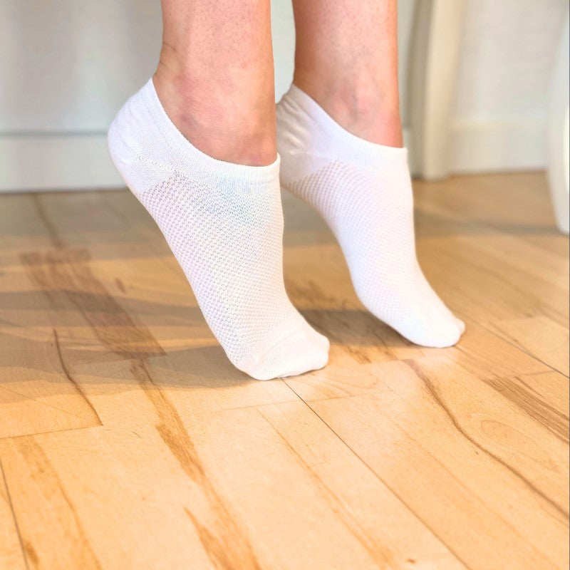 En person står på tæer på et lyst trægulv iført Festival Strømpers hvide, nedringede netsokker med bambusfiber. Kraftig belysning fremhæver de temperaturregulerende egenskaber, sokkens tekstur og træets åremønster.