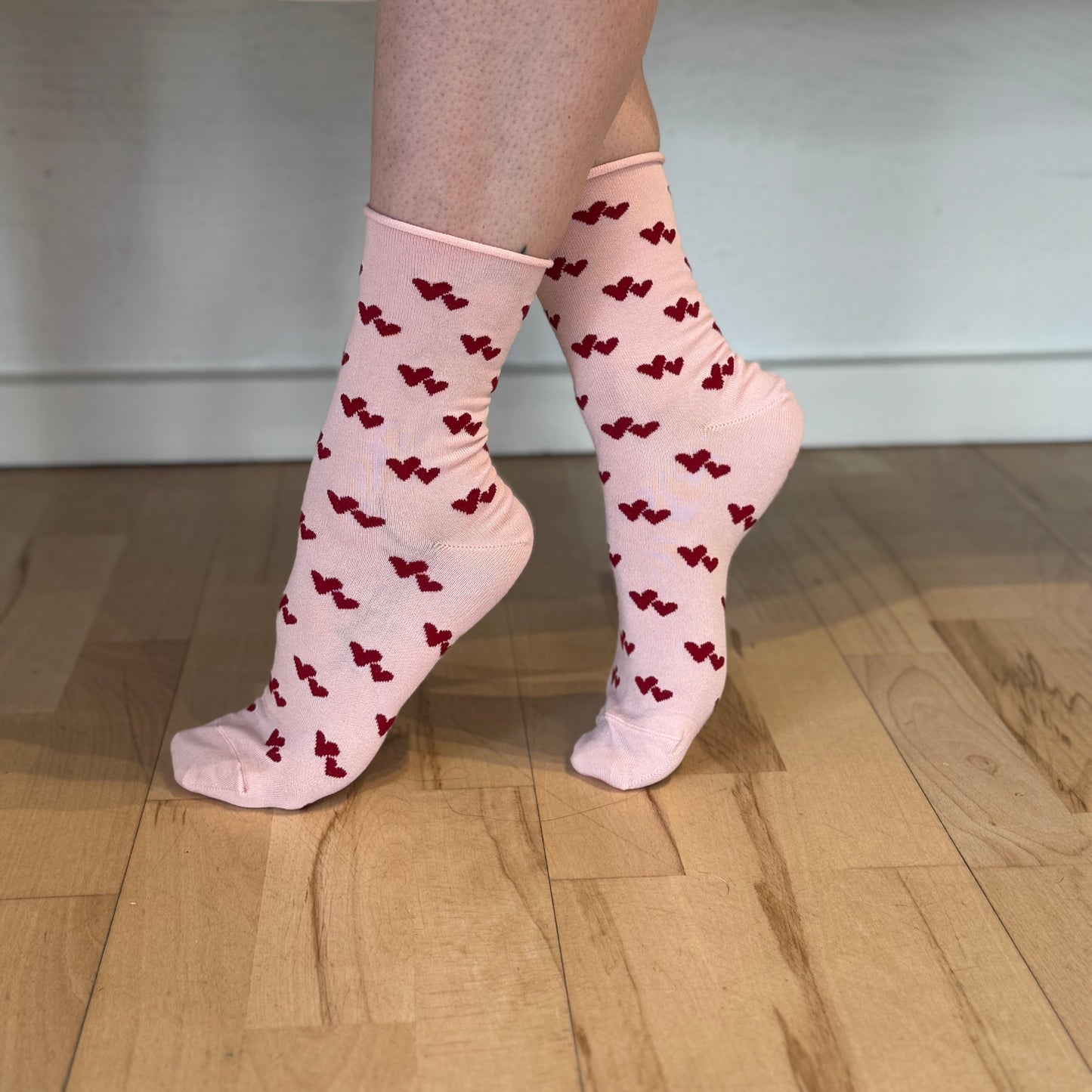 En person balancerer på tåspidserne på et lyst trægulv iført pink Note bambus strømper med små røde hjertemønstre. Kun deres underben og fødder er vist mod en almindelig hvid baggrund.