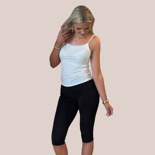 En kvinde med langt blond hår bærer en hvid spaghetti-top og sorte capri-leggings fra Festival Strømper, der symboliserer stil og komfort. Hun ser ned og rører blidt sit hår mod en ensfarvet lys beige baggrund.