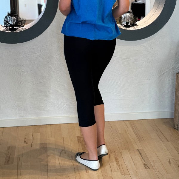 En person står indendørs på et lyst trægulv og kigger væk. De er iført sorte Teyli-leggings for optimal komfort, en lyseblå top og hvide ballerinaer med sort kant. To runde spejle pynter på den moderne hvide strukturerede væg foran.