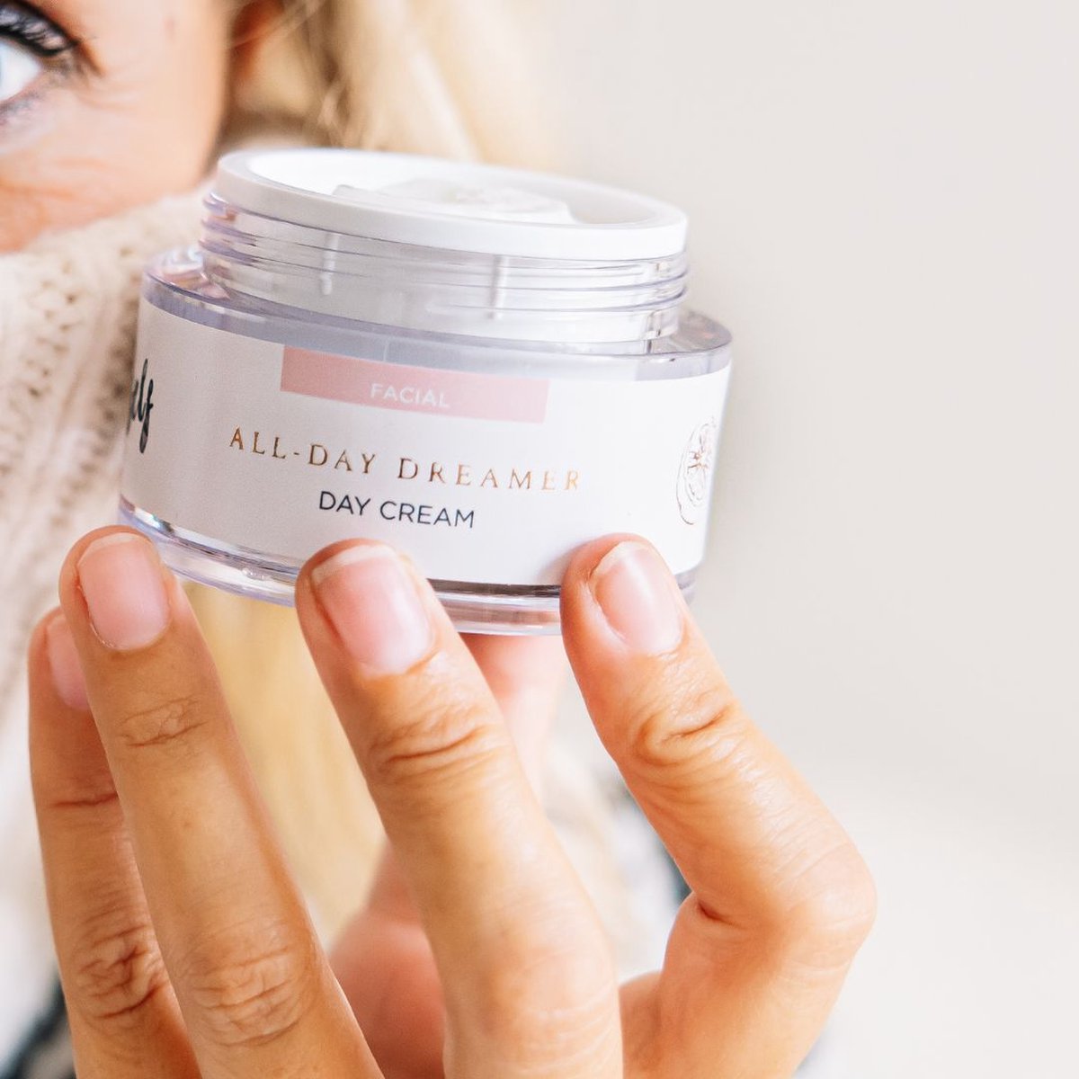 En lyshudet, lyshåret person holder en rund krukke med Natural Self vegansk dagcreme tæt på ansigtet. Den hvide etiket med lyserøde accenter og de velklippede negle skiller sig ud mod en let sløret baggrund.