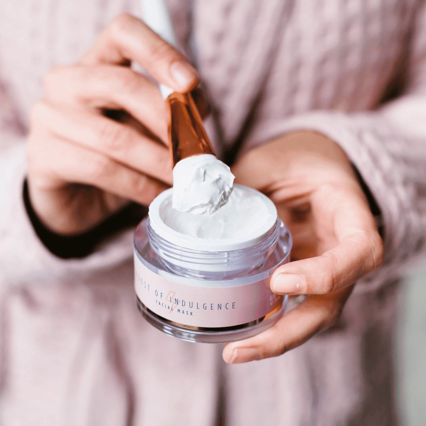 En person i en pink tekstureret morgenkåbe holder en åben krukke med Natural Self Ritual of Indulgence Facial Mask, en anti-age ansigtsmaske, i den ene hånd og øser lidt ud med en børste i den anden.