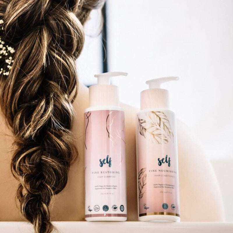 Et nærbillede af en kvindes ryg afslører hendes lange, tykke, brune hår i en løs fletning. Foran står to pumpeflasker med Natural Self vegansk shampoo med pastelfarvede etiketter på en lys overflade i blødt, naturligt lys.