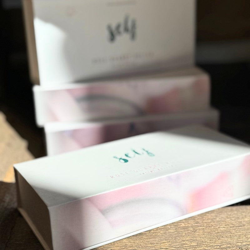 Tre pastelfarvede rektangulære kasser med påskriften "Natural Self" er stablet på en træoverflade, mens sollyset lyser op i toppen. Den beroligende scene indbyder til øjeblikke med selvpleje, perfekt til hudpleje eller til at bruge din foretrukne Rose Quartz Facial Roller.