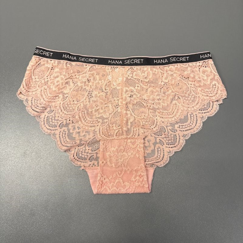 Et par lyserøde JESSYLIA Lace-trusser med skulpterede kanter vises på en grå overflade. Den sorte linning har gentagende hvid HANA SECRET-tekst, og de blomstrede blonder giver et feminint touch til disse rosa trusser.