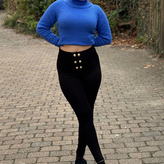 En person står udendørs på en brostensbelagt sti iført en blå rullekravesweater og højtaljede leggings med guldfarvede knapper fra Soy & Co. Armene er bag ryggen, benene er krydsede, og der er grønne planter og et træhegn bag dem.