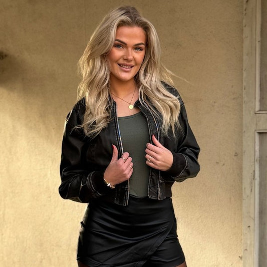 En smilende kvinde med langt blond hår står udenfor iført en Promise kunstlæder bomberjakke over en grøn top og sorte shorts, tilbehøret af en guldhalskæde og armbånd. Hun poserer med den ene hånd, der rører sin jakke op ad en beige væg.