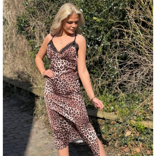 En kvinde med blondt hår står udendørs på en solskinsdag iført en Estee Brown leopardkjole med justerbare stropper. Hun kigger ned og holder let på sin nederdel, der er omgivet af frodigt grønt og en brostensbelagt sti.