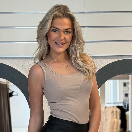 En smilende kvinde med langt, bølget blond hår har en beige Estee Brown-top i behageligt materiale og sorte underdele på. Hun står indendørs op ad en væg med vandrette hvide linjer, bueformede udskæringer og synlige tøjstativer i baggrunden.