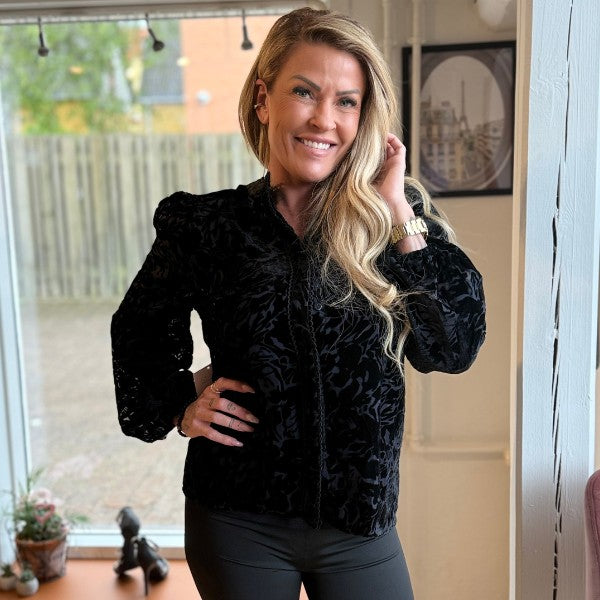 En smilende kvinde med langt blond hår bærer en Zibi London skjorte med fløjsmønster og mørke bukser. Hun står indendørs ved et vindue, rører ved sit hår og har den ene hånd på hoften. I baggrunden ses stueplanter, et træhegn og indrammede kunstværker.