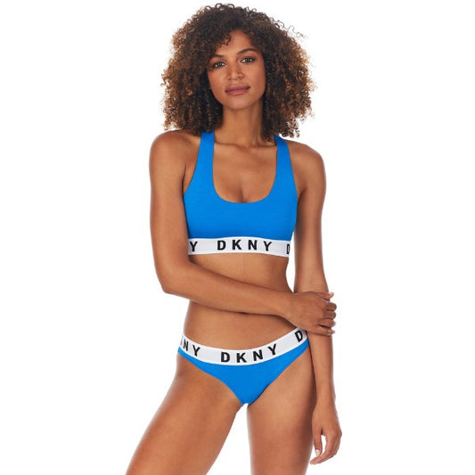 En kvinde med krøllet hår viser en lyseblå DKNY-bralette og matchende trusser, begge med hvide elastiske DKNY-bånd, mens hun poserer selvsikkert mod en almindelig hvid baggrund.