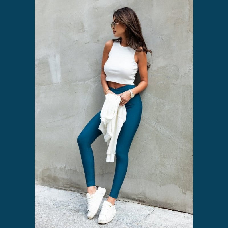 En kvinde poserer afslappet op ad en grå væg i en ærmeløs hvid crop top, Ivon højtaljede blågrønne leggings for komfort og elegance og hvide sneakers, mens hun holder en hvid sweatshirt med solbriller på og sit lange hår løst.