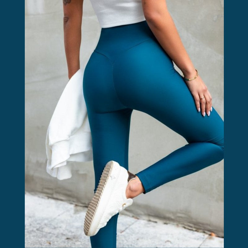 En kvinde står på ét ben iført Ivon's blågrønne højtaljede leggings for komfort og stil, sammen med en hvid ærmeløs top og sneakers. Hun holder en hvid jakke, og hendes lyseblå negle lyser op mod den neutrale væg og stenbelægningen.