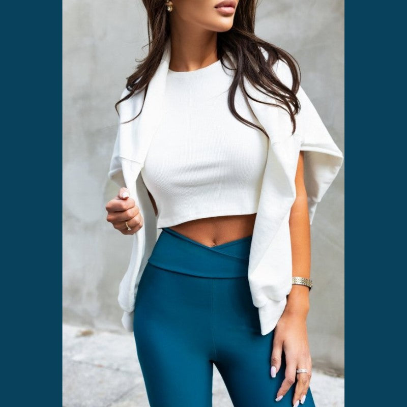 En kvinde med langt brunt hår er iført Ivon høj talje leggings, en hvid crop top og en sweater over skuldrene. Guldøreringe og et ur tilføjer elegance, mens hun står op ad en grå væg og holder sweateren.