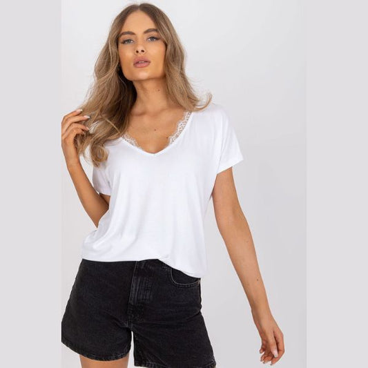 En kvinde med langt, bølget blond hår har en Basic Molly T-shirt i høj kvalitetsbomuld med blondekant og sorte denimshorts med høj talje på. Hun poserer selvsikkert mod en lysegrå baggrund med den ene hånd rørende ved håret.