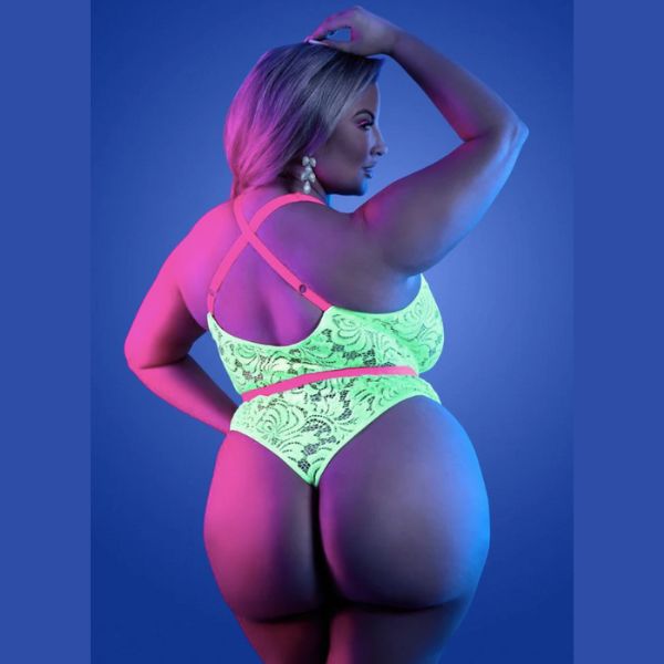 En plus-size kvinde poserer selvsikkert i en neonfarvet teddy med justerbare stropper fra Fantsy Lingerie, vist bagfra mod en blå baggrund. Hun løfter armen over hovedet, mens pink og blå belysning fremhæver hendes kurver.