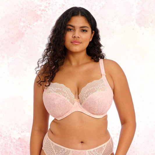 En kvinde med langt, krøllet mørkt hår viser Elomi Lucie Stretch Bh'en i lyserød blonde med matchende undertøj med dyb udskæring og støttende powernet mod en blød lyserød og hvid akvarelbaggrund.