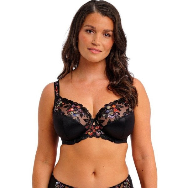 En kvinde med langt, bølget brunt hår modellerer en Fantasie Magdalena Noir sidestøtte-bh med gennemsigtig mesh og blomsterbroderi samt matchende undertøj, mens hun står foran en almindelig hvid baggrund og kigger selvsikkert ind i kameraet.
