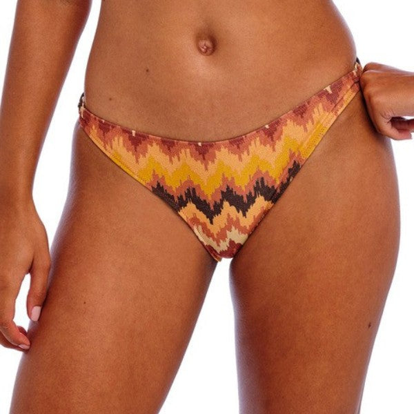 En person viser Freya bikinitrusser med et livligt gult, orange, brunt og sort print. Billedet viser underkroppen og overlårene; den ene hånd hviler på hoften, mens den anden blidt trækker i bikiniens side.