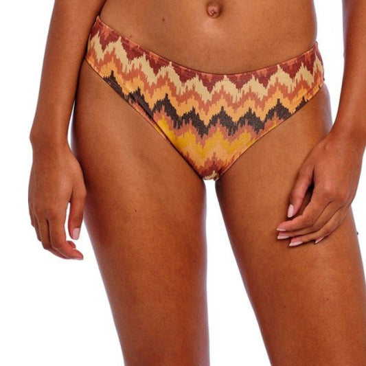 En kvinde med mellembrun hud står mod en hvid baggrund iført Freya bikinitrusser i et Arizona-print i brunt, guld og rødbrunt. Kun hendes torso og overlår er synlige, og hendes arme hviler afslappet langs siderne.