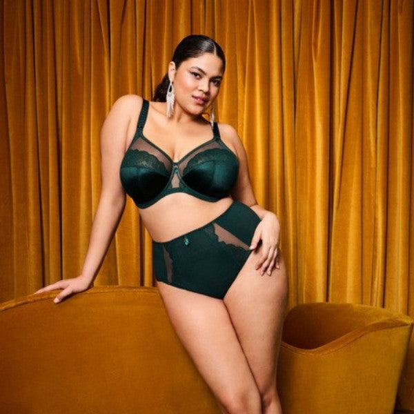 En kvinde med mørkt hår i en glat hestehale poserer selvsikkert i en Elomi smaragdgrøn bøjle-bh med gennemsigtige paneler, stående op ad et guldgardin og lænet op ad en matchende fløjlssofa. Hun bærer statement-øreringe og kigger direkte ind i kameraet.