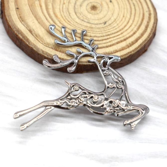 En sølvfarvet broche fra Rouge Bonbons med et springende rensdyrmotiv og elegante snirkler ligger på en hvid overflade ved siden af en rund træskive med synlige årringe.