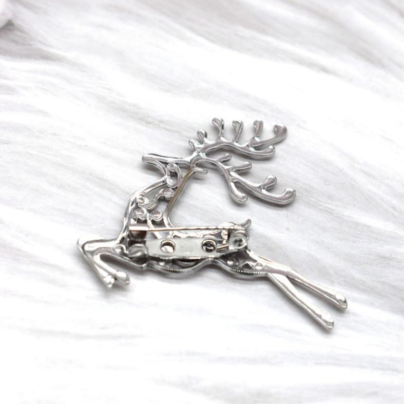 A shiny sølvfarvet Rouge Bonbons broche formet som en løbende hjort med store gevirer ligger på hvid, tekstureret stofbaggrund. Bagsiden med nålemekanisme og lås ses tydeligt.