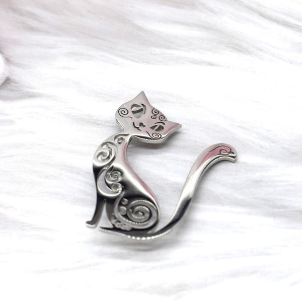 En katteformet sølvbroche fra Rouge Bonbons med hvirvlende mønstre og udskårne øjne og bryst hviler på en blød, hvid pelsoverflade. Dette elegante tilbehør har et minimalistisk kattemotiv design.