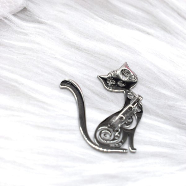 Rouge Bonbons sølvbroche har et elegant kattemotiv i profil med en yndefuldt krøllet hale, detaljeret med indviklede hvirvlende udskæringer. På en blød, hvid pelslignende baggrund tilføjer dette dekorative tilbehør stilfuld charme.