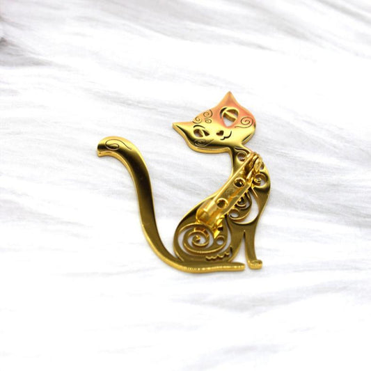 En guldfarvet broche fra Rouge Bonbons formet som en kat med indviklede swirl-designs præsenteret på hvid, tekstureret baggrund. Kattens buede hale og let skrå hoved giver et elegant, kunstnerisk udtryk.