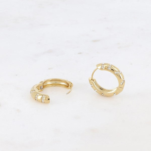 To små guldøreringe fra Fashion Paris ligger på en hvid flade. Hver har små zirkoniumkrystaller i klynger langs kanten for et delikat, elegant look. Den ene ørering er lukket, mens den anden forbliver åben.