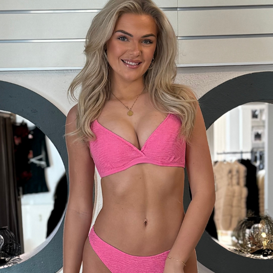 En smilende kvinde med langt blond hår iført en lyserød Freya-bikini står indendørs, omgivet af cirkulære spejle, en grå og hvid væg og tøjstativer – et perfekt sommergarderobe-valg til et trendy strandlook.