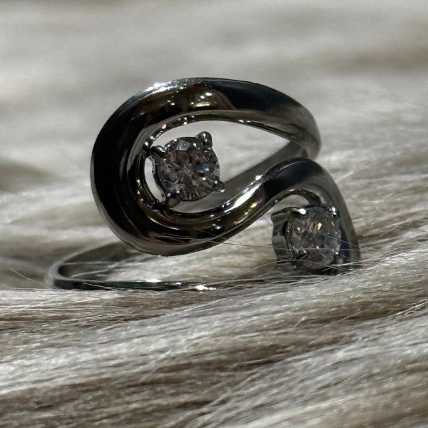 Et nærbillede af en justerbar Rouge Bonbons-ring i rustfri stål med et moderne, snoet design og to klare runde ædelsten sat overfor hinanden, vist på blødt, lyst pelslignende materiale.