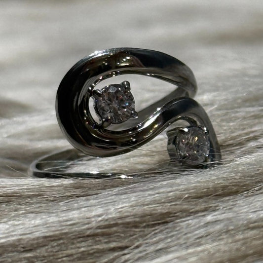 Et nærbillede af en justerbar Rouge Bonbons-ring i rustfri stål med et moderne, snoet design og to klare runde ædelsten sat overfor hinanden, vist på blødt, lyst pelslignende materiale.