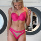 En blond kvinde modellerer en hot pink Axami bh og et brasiliansk trussæt med gennemsigtige paneler, mens hun står indendørs ved en hvid væg med to store runde spejle og pynter sig med smykker.