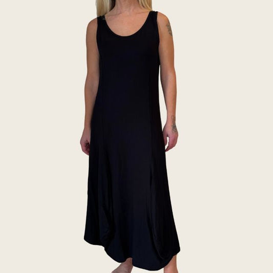 En kvinde med langt blondt hår bærer en sort, ankellang sommerkjole fra E Avantgarde lavet af økologisk viskose. Hun står barfodet på en almindelig, lys baggrund med armene afslappet langs siden og ansigtet ude af syne.