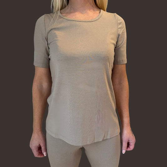 En kvinde med lys hud og glat blondt hår bærer en tætsiddende, kortærmet sandfarvet T-shirt og leggings fra E Avantgarde. Hun står med ansigtet fremad mod en mørkebrun baggrund med armene langs siden og ansigtet skjult.
