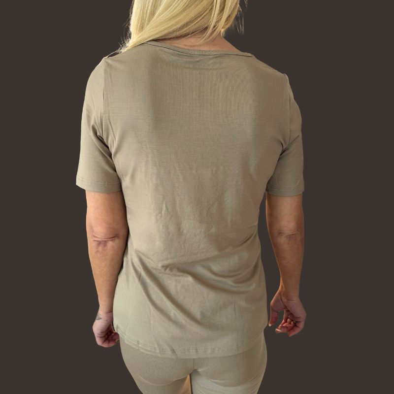 En person med langt blondt hår, set bagfra, er iført en kortærmet E Avantgarde T-shirt i sandfarvet og matchende bukser. Baggrunden er ensfarvet mørk, og deres arme hviler afslappet i siden.