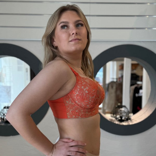 En kvinde med langt blond hår i profil bærer en orange blondebralette fra Empreinte, der udstråler fransk elegance. Hun ser opad med et neutralt udtryk, modsat hvide vandrette linjer og to store runde spejle, der reflekterer lys.