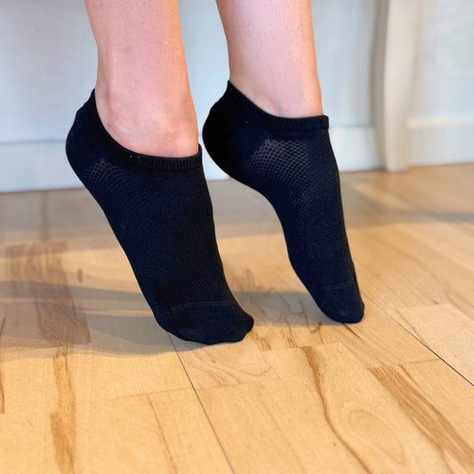 En person står på tæer på et lyst trægulv iført sorte Festival Strømper ankelsokker med bambusfiber til effektiv svedtransport. I baggrunden er en hvid væg og en del af et lyst møbelben til højre.