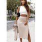 En kvinde står udendørs i klassisk stil iført en hvid crop top fra Ivon og en beige midi-nederdel med slids i siden. Hun har en matchende blazer, bærer en sort skuldertaske med guldkæde og pynter sig med guldsmykker.