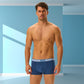 En veltrænet mand med kort krøllet hår står i et blåt rum iført Henderson-boxershorts med en hvid HENDERSON-linned, mens geometriske former kaster skygger på væggene.